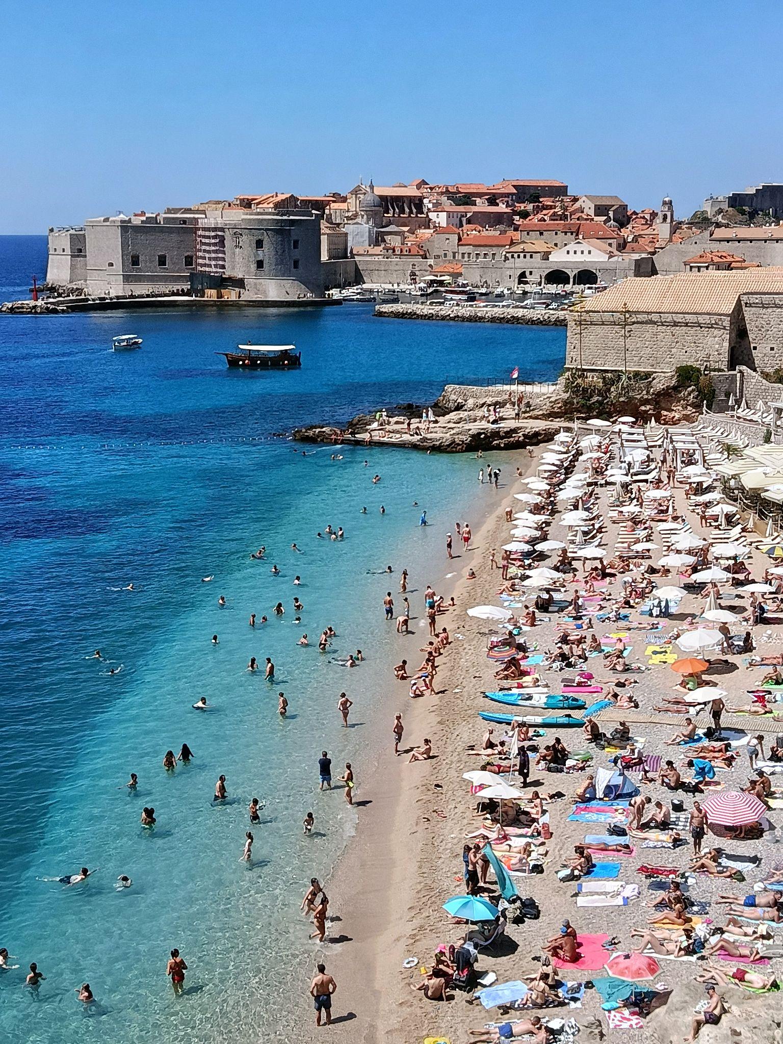 Banje Beach in Dubrovnik with swimmers in clear turquoise water and sunbathers on the sandy shore, overlooking the Old Town walls.