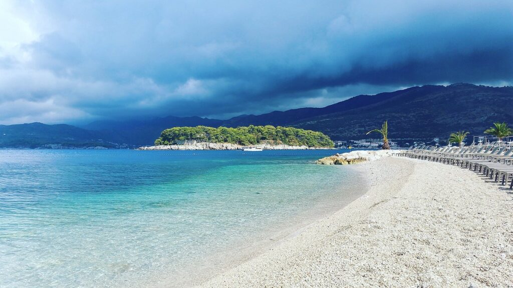 Copacabana Beach Dubrovnik with white pebble shore, calm turquoise Adriatic Sea, and view of Daksa Island under dramatic clouds