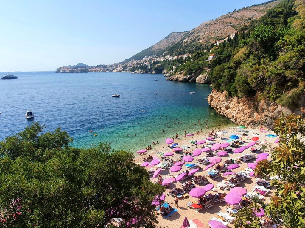 Sveti Jakov Beach Dubrovnik with pink umbrellas, clear turquoise sea, and scenic Old Town coastline view from the cliffs