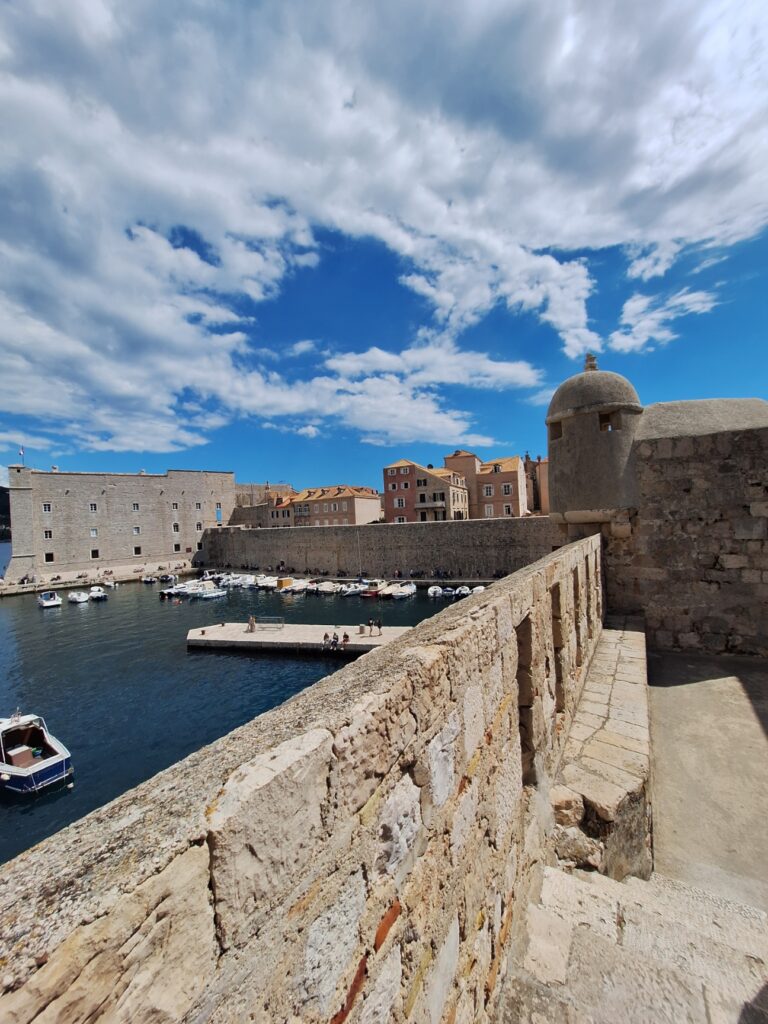 View from the Walls of Dubrovnik overlooking Dubrovnik Old Port, historic stone fortifications, Adriatic Sea, and medieval architecture under dramatic clouds.