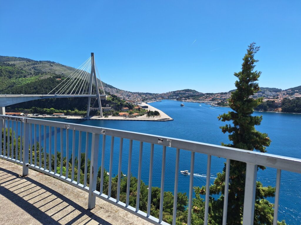 Panoramic view of Franjo Tuđman Bridge overlooking Gruž Harbor, the Adriatic Sea, and the surrounding hills of Dubrovnik on a clear sunny day.