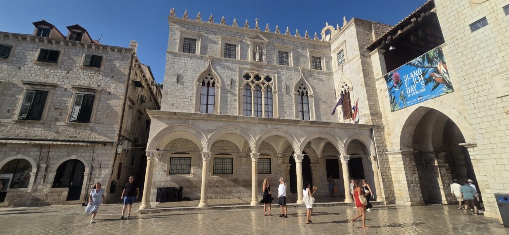 Sponza Palace in Dubrovnik Old Town during the afternoon, with sunlit stone arches, bright square and visitors walking nearby
