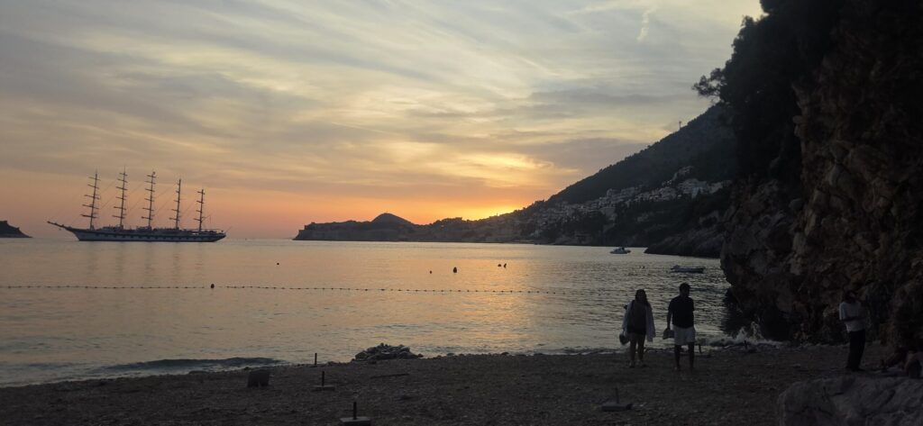 Couple walking along Sveti Jakov Beach at sunset with Dubrovnik and a sailing ship in the distance