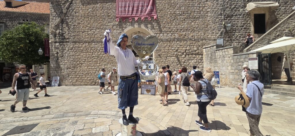 Mirso holding the Dubrovnik Heart souvenir near Pile Gate in Dubrovnik Old Town, with visitors and the historic stone walls behind him.