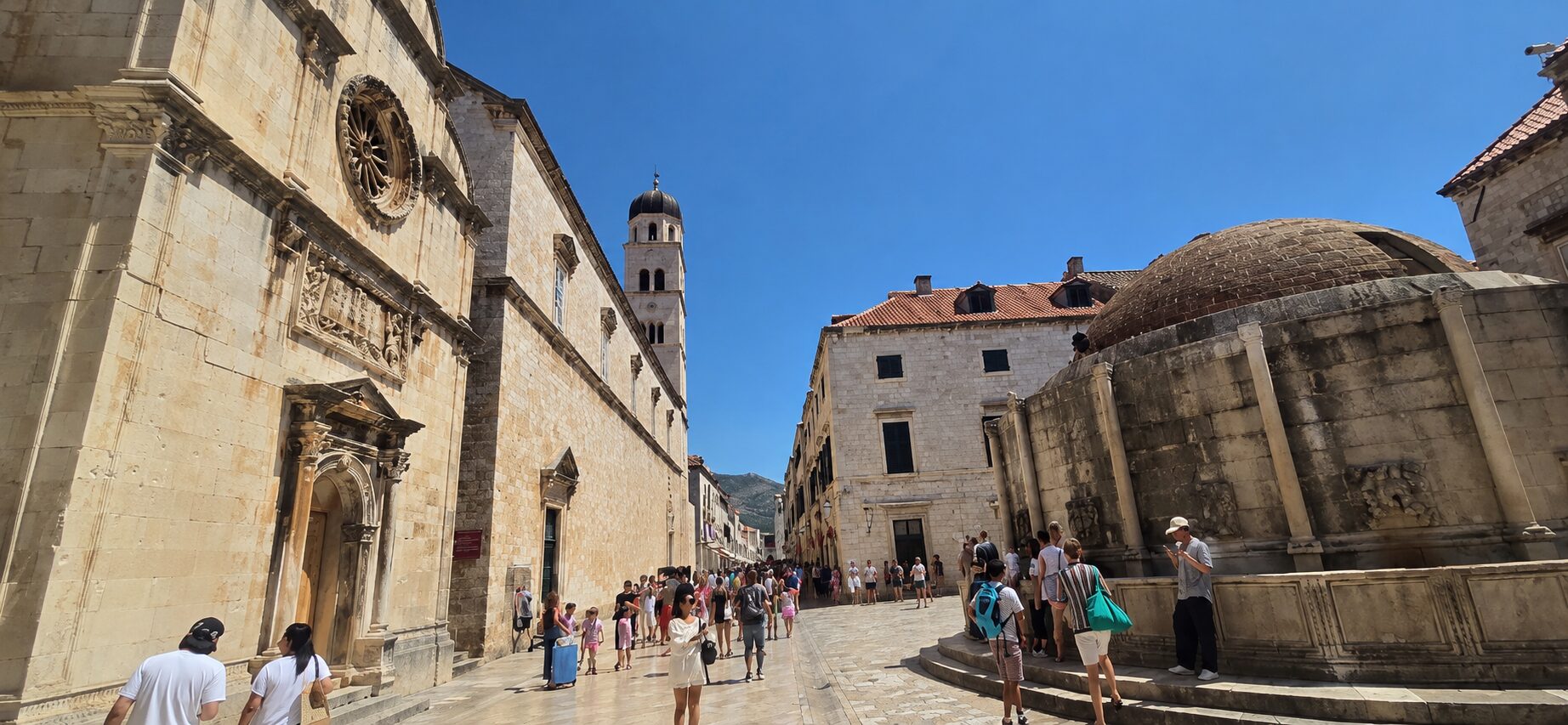 Stradun in Dubrovnik Old Town with the Franciscan Church, Large Onofrio’s Fountain and Bell Tower under a clear summer sky.